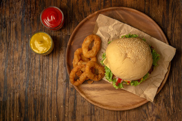 Top view of cheese burger with onion rings on round wooden plate and ketchup and mustard sauce. 