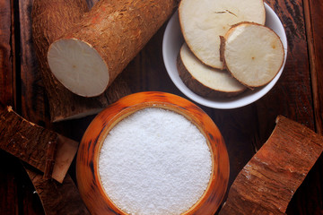 tapioca flour in wooden bowl on rustic wooden table, next to cut and sliced cassava