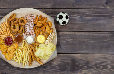 Diverse snacks and beer with a soccer ball on a beer foam on dark wooden background. Top view. Empty space for text