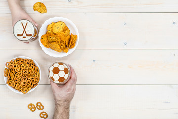 Couple holds glass mug of beer with silhouettes of hockey sticks and soccer ball on beer foam and pretzels with chips on light wooden background. Empty space for text. Top view