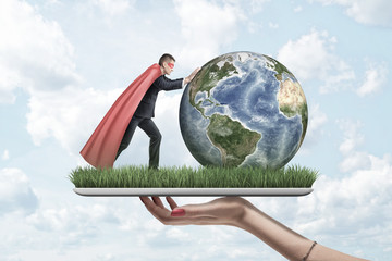 Side view of businessman in red cape and black mask pushing Earth forward, both on digital tablet covered with grass, held by woman's hand.