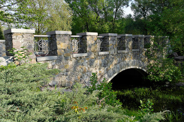 Stone bridge over the creek. flowing among large green trees and bushes. Early summer morning.