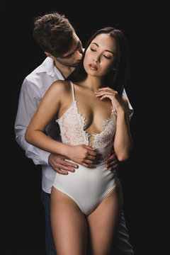 handsome young man embracing and kissing attractive asian girlfriend in white lingerie isolated on black
