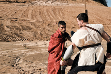 A concept photo of a Roman battle of two warriors of the Colosseum in action with aggressive emotions in full military uniform on a desert landscape on a sunny day with a dry sun.