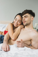 Obraz na płótnie Canvas happy asian woman embracing pensive shirtless boyfriend in bedroom