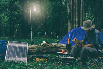 Young caucasian man writing on his note sitting on a camping chair in the woods. Power on light  from the solar battery.