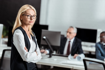 selective focus of attractive businesswoman in glasses standing with crossed arms near multicultural men
