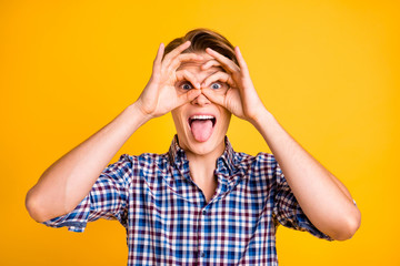 Close up photo amazing youth he him his man hand arm finger okey symbol near eyes specs shape tongue out mouth youngster wear casual plaid checkered shirt outfit isolated yellow bright background