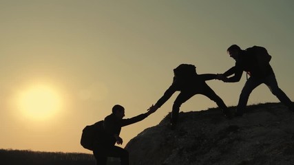 Silhouettes of tourists climbing mountain cliffs against the backdrop of a sunset, helping each other's hand. Help in the mountains and teamwork in hiking. Teamwork concept.