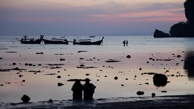 silhouette of a couple sitting on a beach, with long tail boats in the background with people walking through water in sunset on the beach. Thailand. Ton Sai beach