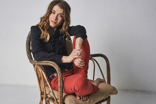 young, sympathetic, emotional, sexy, European light girl with loose hair in red jeans and a black shirt sits in a wicker armchair. On a light background - Powered by Adobe