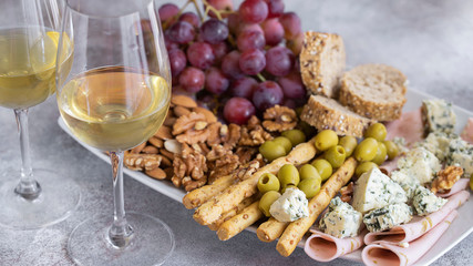 Two glasses of white wine and plate with different snacks. Blue cheese, olives, baguette slices, grissini, ham, grapes and nuts. Wine snacks set background.