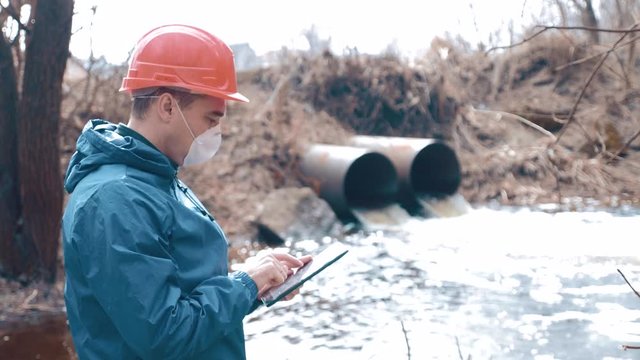 Environmental pollution. A man in a helmet and respirator with a tablet measures the level of pollution of waste water from a pipe. Environmental conservation concept