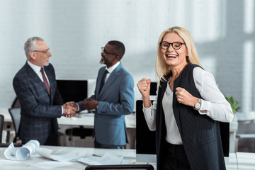 selective focus of happy businesswoman celebrating triumph near multicultural businessmen shaking hands in office