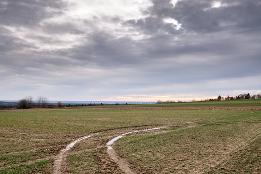Moody weather with grey clouds in the sky above an agricultural field with a little green an puddles in the tractor tracks. Seen in March 2019 near Heroldsberg, Germany