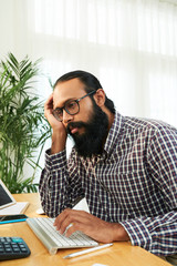 Tired bearded businessman in eyeglasses working at the table on computer keyboard at office