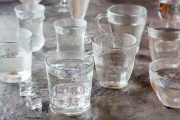 Many glasses of water splashed with drops and pieces of ice on a gray table