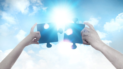 Two hands holding a puzzle pieces agains of blue sky background and sun. Business, support, finding...