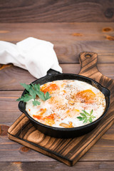 Homemade breakfast of shakshuka fried eggs with tomatoes and herbs in a pan on a wooden table