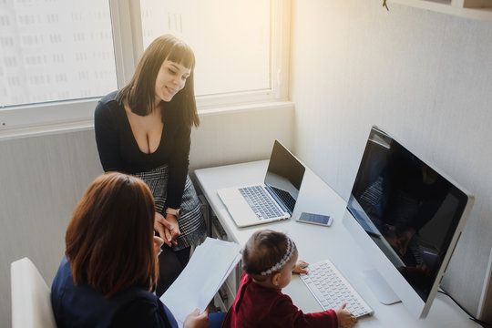 close up photo of two women in office outfit working on computers and discussing a report on the floor; one of them is with a baby girl in her arms