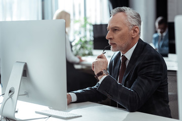 selective focus of pensive mature businessman holding glasses and looking at computer monitor near multicultural colleagues in office