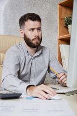 Fototapeta premium Bearded serious manager concentrating on his work on computer while sitting at workplace at office