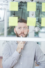 Fototapeta premium Pensive young businessman standing near the glass wall looking at graph and thinking over his business plan