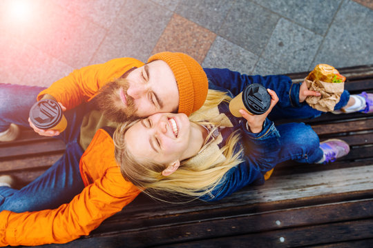 Stylish hipster couple laughing, joking spending time together outdoors. Top view of attractive woman and handsome bearded man are sitting back to back on a bench with coffee cupin the city street