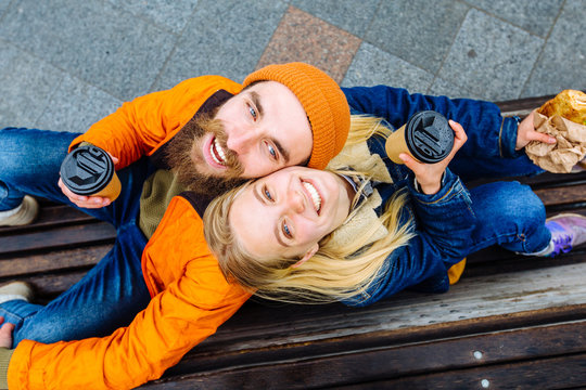 Stylish hipster couple laughing, joking spending time together outdoors. Top view of attractive woman and handsome bearded man are sitting back to back on a bench with coffee cupin the city street