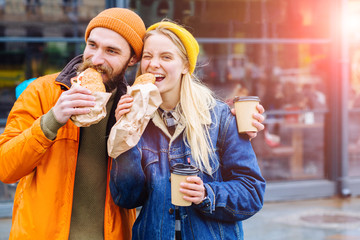 Couple is hungry and eating a burger croissants at lunch time in the city street outdoor. Beard man...