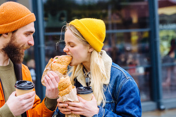 Funny beard hipster man feeding blond hungry caucasian woman with croissant or sandwich outdoors in...