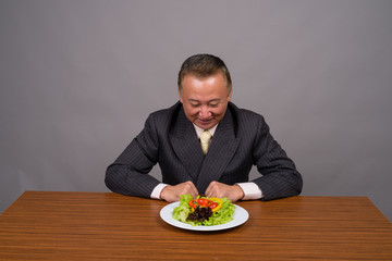 Mature Asian businessman sitting with wooden table against gray 