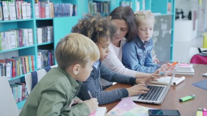 Tracking right shot of female teacher and three elementary students sitting at desk in school library and doing tasks on laptop computer together