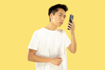 Korean young man's half-length portrait on yellow studio background. Male model in white shirt. Using smartphone. Concept of human emotions, facial expression. Front view. Trendy colors.