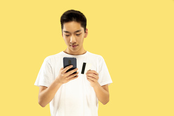 Korean young man's half-length portrait on yellow studio background. Male model in white shirt. Paying bill or buying online. Concept of human emotions, facial expression. Front view. Trendy colors.