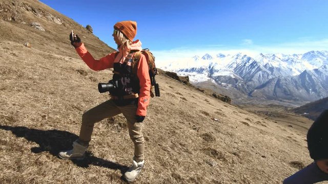 Portrait of a happy girl traveler photographer in a hat and sunglasses with a camera around his neck and a phone in his hands on the background of mountains takes pictures on his smartphone