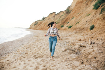 Naklejka premium Stylish hipster girl running on beach at sea and smiling. Happy boho woman in denim jeans and floral blouse relaxing at sandy cliff on tropical island. Travel and summer vacation concept