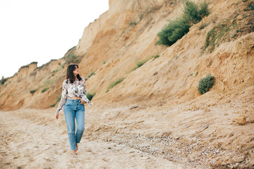 Naklejka premium Stylish hipster girl walking on beach at sea and relaxing. Happy boho woman in denim jeans and floral blouse relaxing at sandy cliff on tropical island. Travel and summer vacation concept