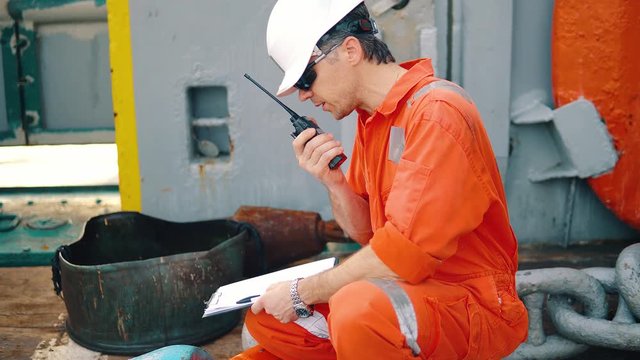 Marine chief officer or chief mate on deck of ship or vessel. He fills up ahts vessel checklist. Ship routine paperwork. He holds VHF walkie-talkie radio in hands.