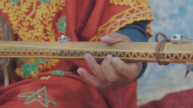 Close up of a Moroccan mans hands playing a old style musical instrument white dressed in traditional orange clothing while sitting on the street