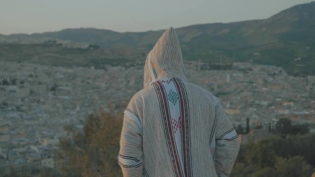 Panning around a man in a cream colored jumper with hood facing away from the camera arms open on a mountain top overlooking city of Fez, Morocco with hundreds of stone houses and buildings below