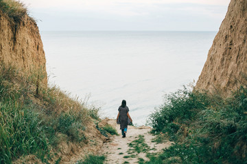 Stylish hipster girl walking to sea between sandy cliffs with grass at sunset. Happy young boho woman exploring tropical island at cliff beach in evening. Summer vacation. Space for text.
