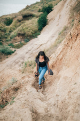 Naklejka premium Stylish hipster girl relaxing on sandy cliff and holding photo camera. Happy young boho woman exploring tropical island at sandy cliff beach and grass. Summer vacation. Space for text.