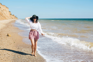 Fototapeta premium Happy young boho woman walking in sea waves in sunny warm day at tropical island and blue sky. Space for text. Stylish hipster girl in hat relaxing on beach and smiling. Summer vacation