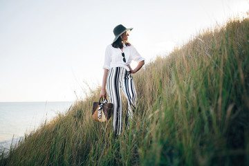 Stylish boho girl in hat posing at sandy cliff with grass near sea, in sunny light. Happy young hipster woman relaxing on tropical island beach in evening. Summer vacation. Space for text