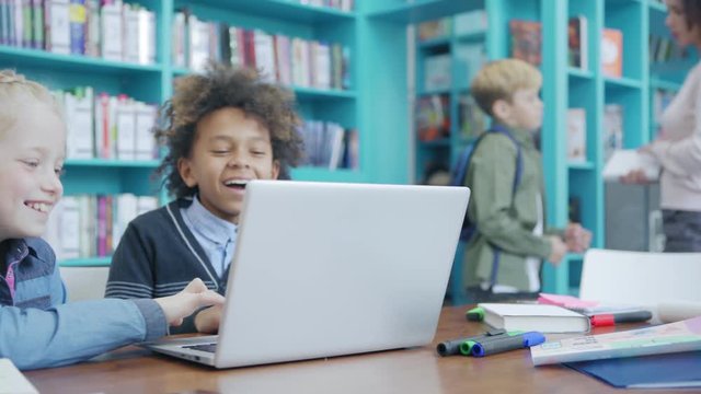 Tracking shot of two diverse elementary school kids, Caucasian girl and mixed race boy, laughing while enjoying playing educational games on laptop computer, teacher talking to student in background