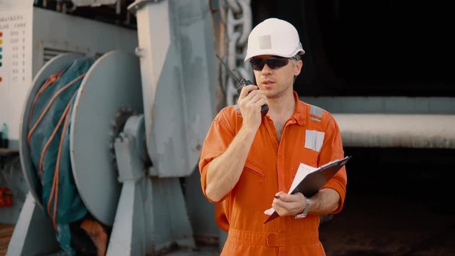 Marine chief officer or chief mate on deck of ship or vessel. He fills up ahts vessel checklist. Ship routine paperwork. He holds VHF walkie-talkie radio in hands.