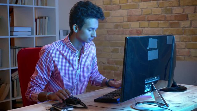 Closeup shoot of young indian attractive male programmer typing on the computer sitting indoors in the cozy apartment with neon light
