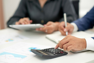 Fototapeta premium Close-up of businessman sitting at office desk with financial graphs and calculating incomes of the company with businesswoman using tablet pc near by him
