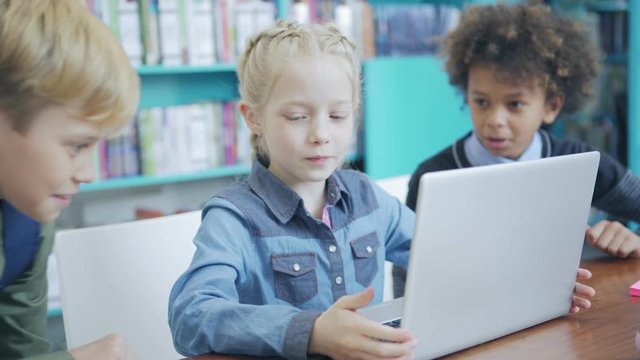 Cute schoolgirl sitting at desk and doing task on laptop computer. Two boys joining her in elementary school classroom to help and advise